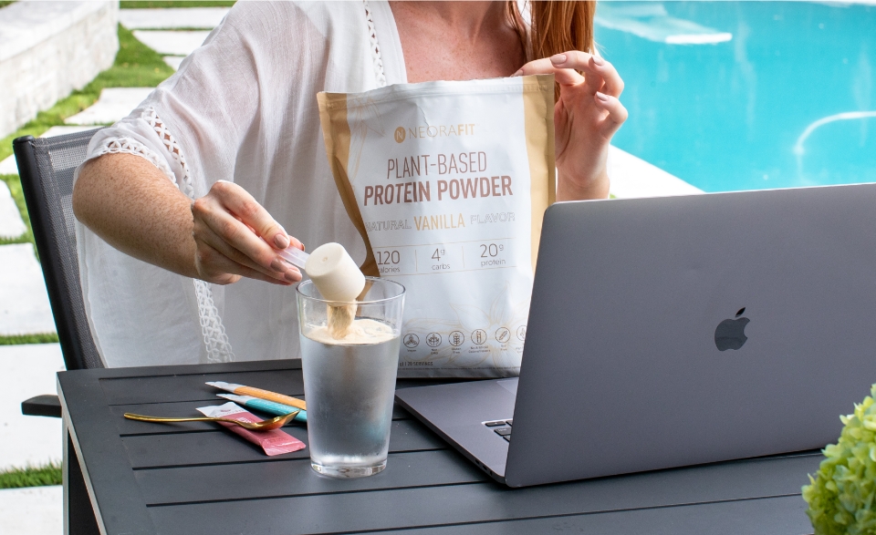A woman pouring Plant-Based Protein Powder into a glass cup outside next to a swimming pool.
